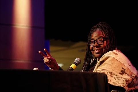 Loretta J. Ross seated at a black table, speaking, holding two fingers in the air