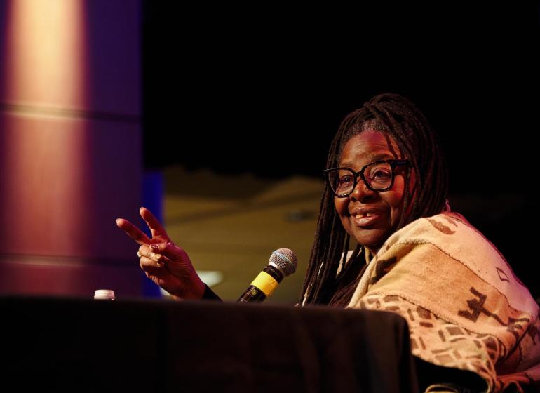 Loretta J. Ross seated at a black table, speaking, holding two fingers in the air