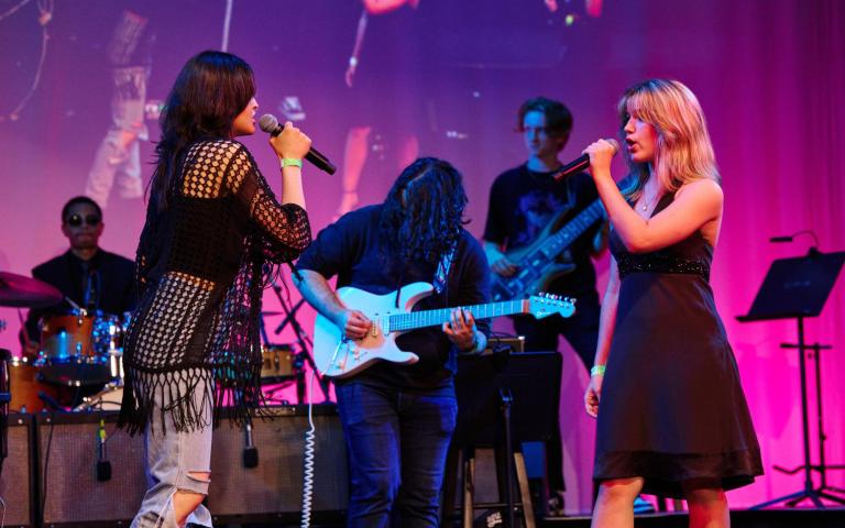 Two young women - one brunette and one blonde, face each other with microphones in their hands on the Berklee Performance Center stage. A guitarist with long dark hair stands in the middle of them.