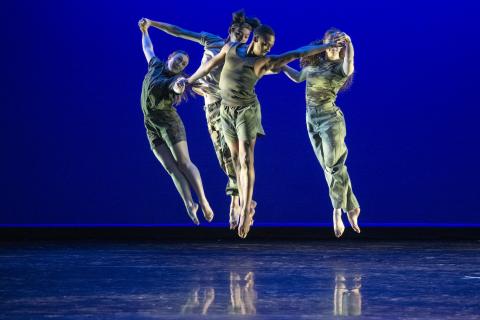 Photo of four student dancers holding hands while jumping in the air in unison against a royal blue background.
