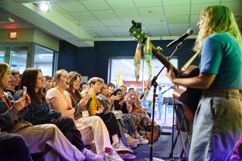 Student sings and plays guitar and other students in the audience smile and take pictures