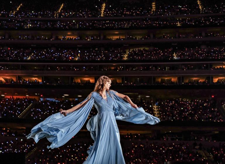 Taylor Swift in a flowy dress on stage with a backdrop of a packed stadium of fans