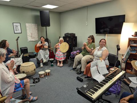 A circle of women seated, playing piano and percussion together