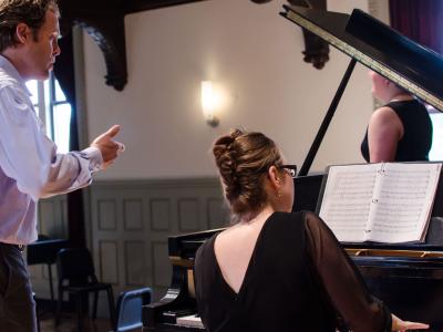 Man standing behind woman who is seated at a piano accompanying a vocalist