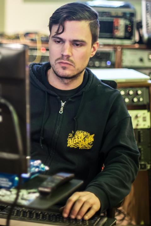 Benny Grotto sitting and looking at a computer in his recording studio, surrounded by gear