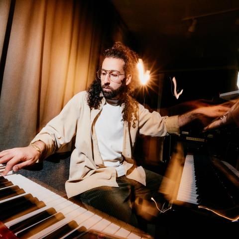 Photo of Yessaï Karapetian playing a piano and keyboard with each hand. He has long curly black hair, a beard, and glasses.
