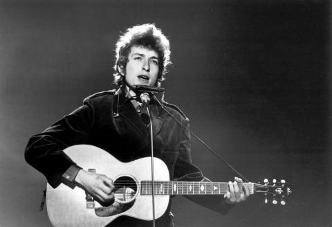Black and white image of a young Bob Dylan performing on stage with an acoustic guitar and harmonica