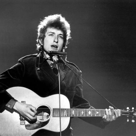 Black and white image of a young Bob Dylan performing on stage with an acoustic guitar and harmonica