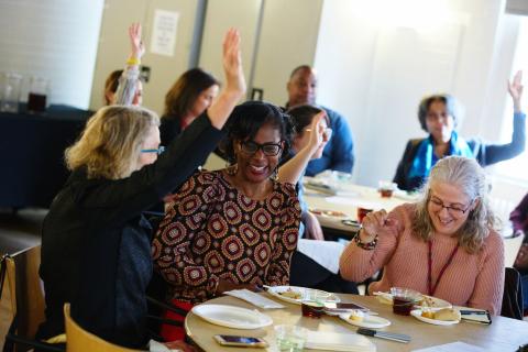 Faculty and staff sitting at a table participate in a discussion and raise their hands