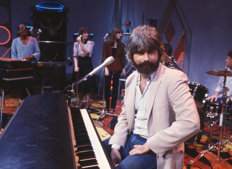 A young Michael McDonald sitting on stage at a piano with his band in the background
