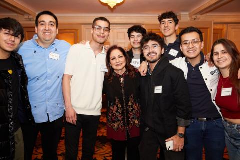 A group of nine people, consisting of students and Gloria Stefan, pose together for a photo indoors, all smiling and wearing casual to semi-formal attire with name tags visible.
