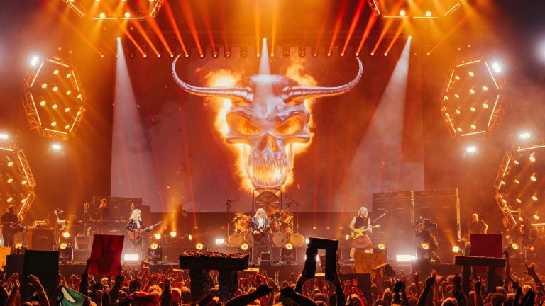 Spinal Tap on stage from a crowd perspective, with fans holding up miniature stonehenges and a giant flaming skull behind the band
