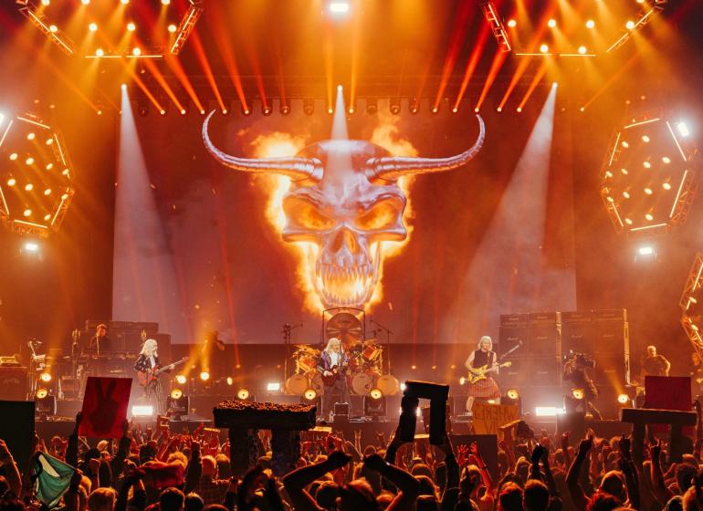 Spinal Tap on stage from a crowd perspective, with fans holding up miniature stonehenges and a giant flaming skull behind the band