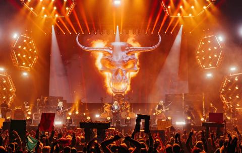 Spinal Tap on stage from a crowd perspective, with fans holding up miniature stonehenges and a giant flaming skull behind the band