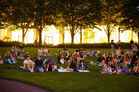 Students sit on a lawn with trees in the background