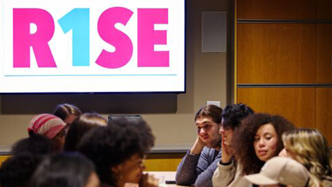Students seated around a table together with a poster behind them that reads "R1SE"