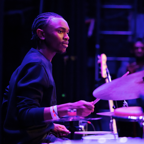 Student plays drum set wearing a black sweater and braids. 